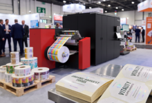 Impressora digital de rótulos Ecolyne, da Flint Group Digital Xeikon, em operação em estande de feira do setor, imprimindo rótulos coloridos para embalagens de alimentos e suplementos nutricionais, com rolos de rótulos acabados em primeiro plano.