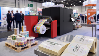 Impressora digital de rótulos Ecolyne, da Flint Group Digital Xeikon, em operação em estande de feira do setor, imprimindo rótulos coloridos para embalagens de alimentos e suplementos nutricionais, com rolos de rótulos acabados em primeiro plano.